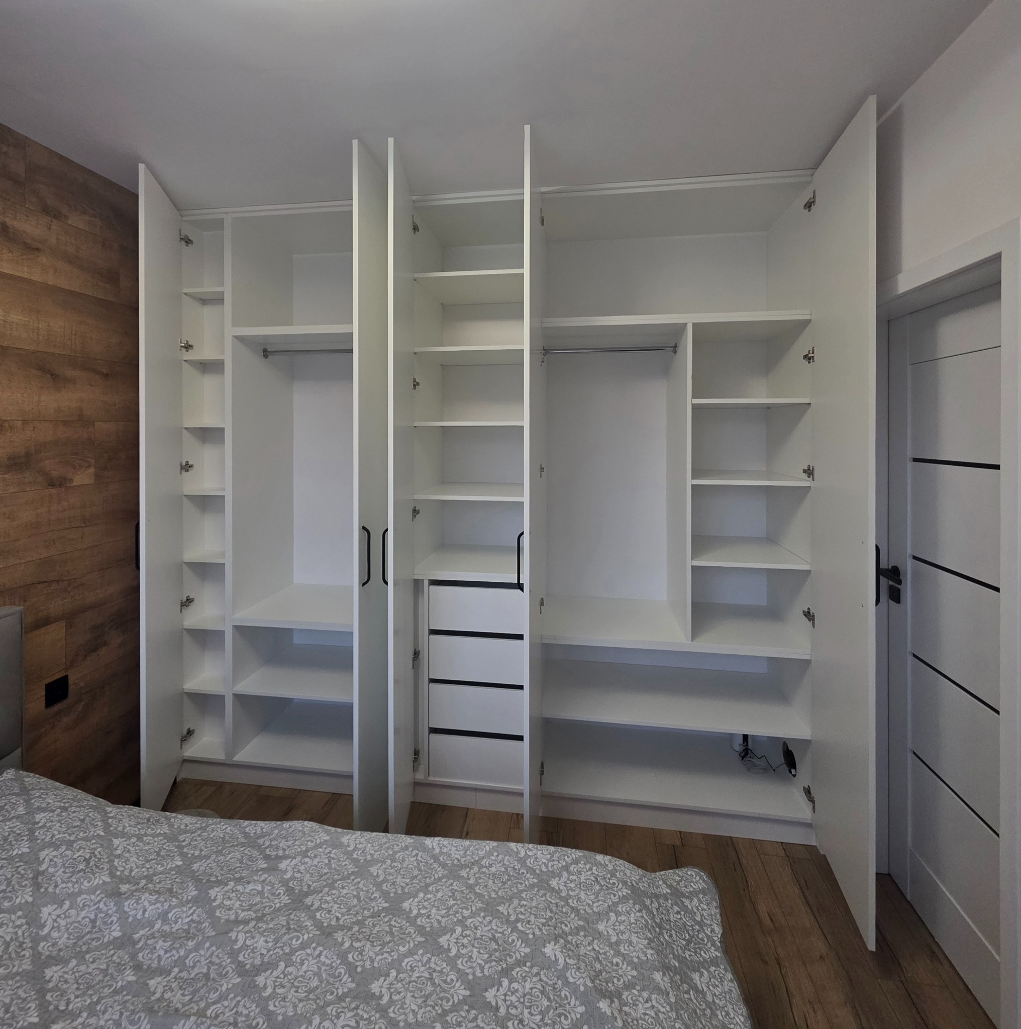 Biała szafa wnękowa z otwartymi drzwiami, widoczne półki, drążki na ubrania i szuflady, w tle drewnopodobna ściana i podłoga, oraz częściowo widoczne łóżko z wzorzystą pościelą.