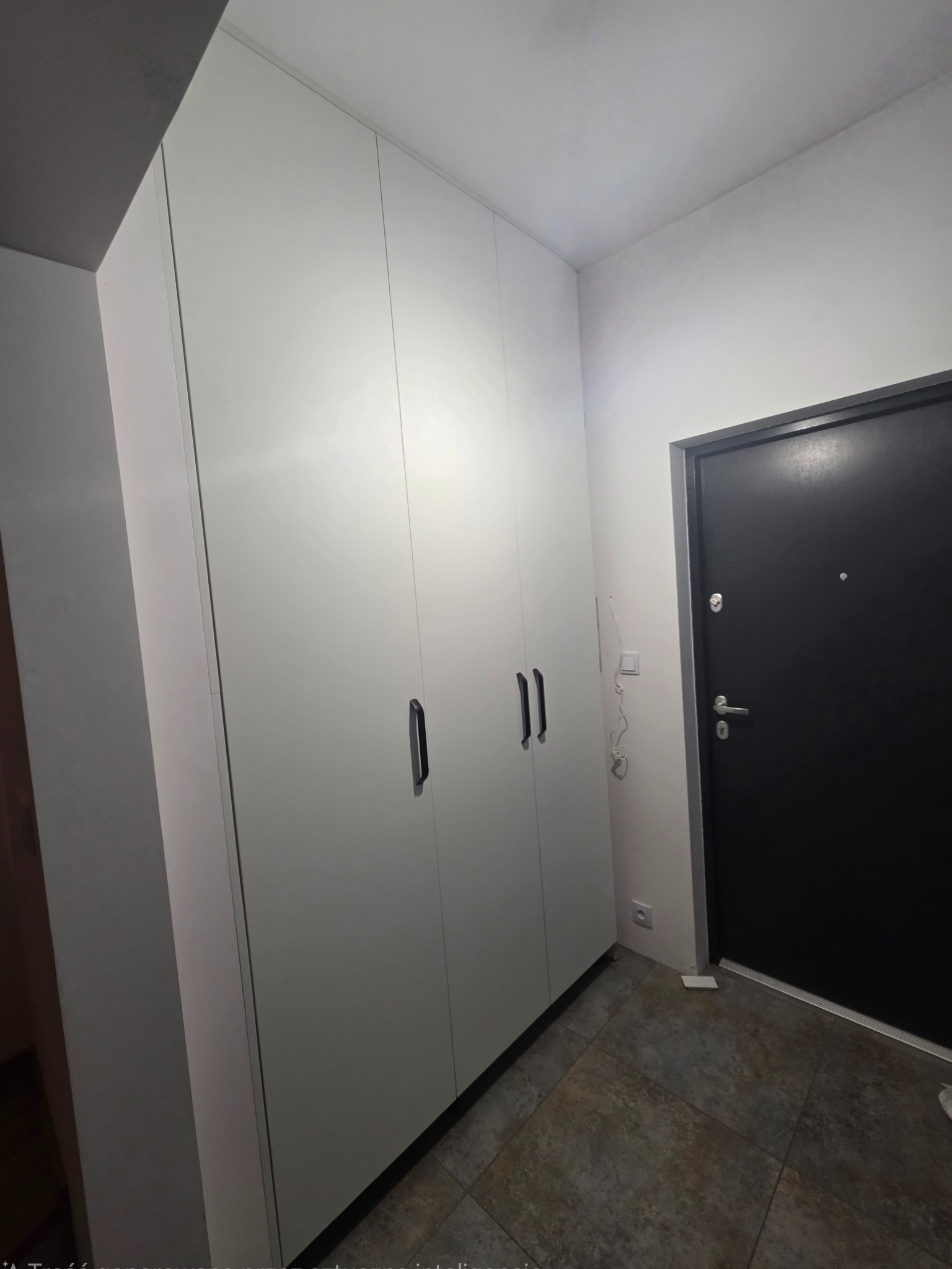 Biała szafa wnękowa z trzema drzwiami i czarnymi uchwytami w przedpokoju z ciemną podłogą i czarnymi drzwiami wejściowymi.