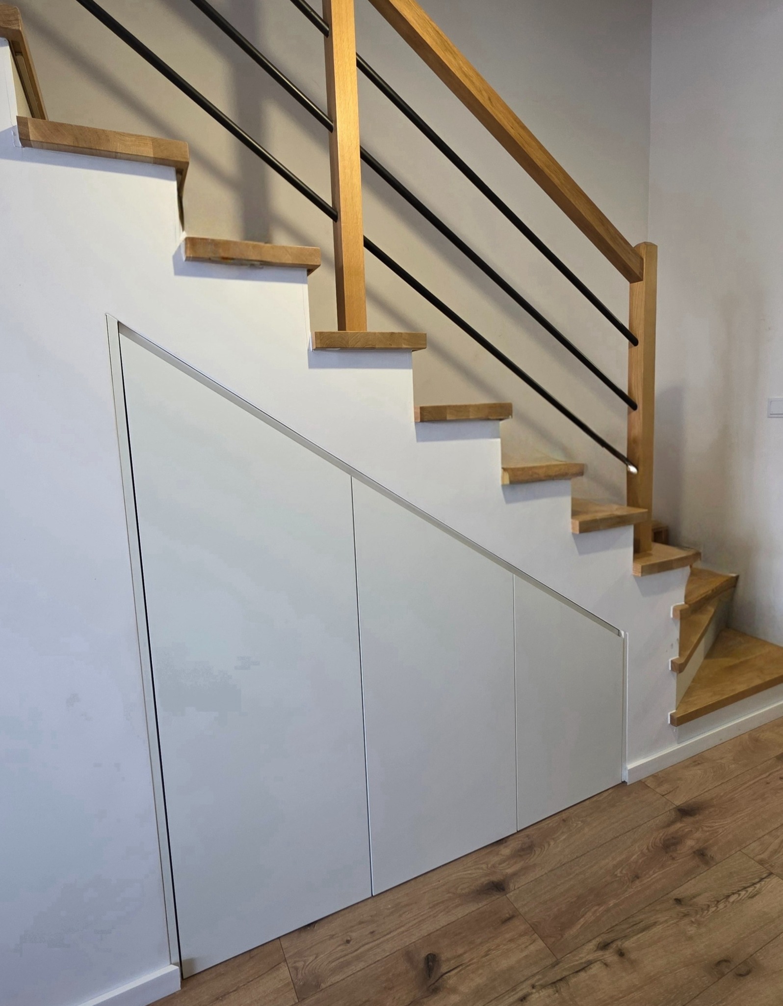 Biała zabudowa szafy pod schodami z drewnianymi stopniami i czarną balustradą, widok na dolną część schodów i podłogę z paneli.