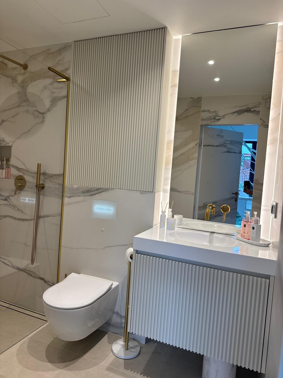 Nowoczesna łazienka z szafką umywalkową z ryflowanym frontem, marmurowym wykończeniem i złotymi akcentami. Kabina prysznicowa ze złotym wykończeniem.