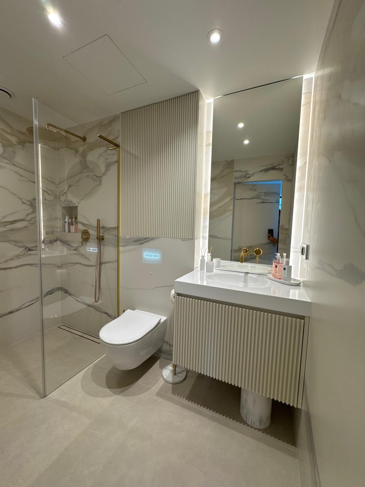 Nowoczesna łazienka z kabiną prysznicową typu walk-in, umywalką z szafką z lamelami i podświetlanym lustrem. Marmurowe wykończenie ścian i złote akcenty.