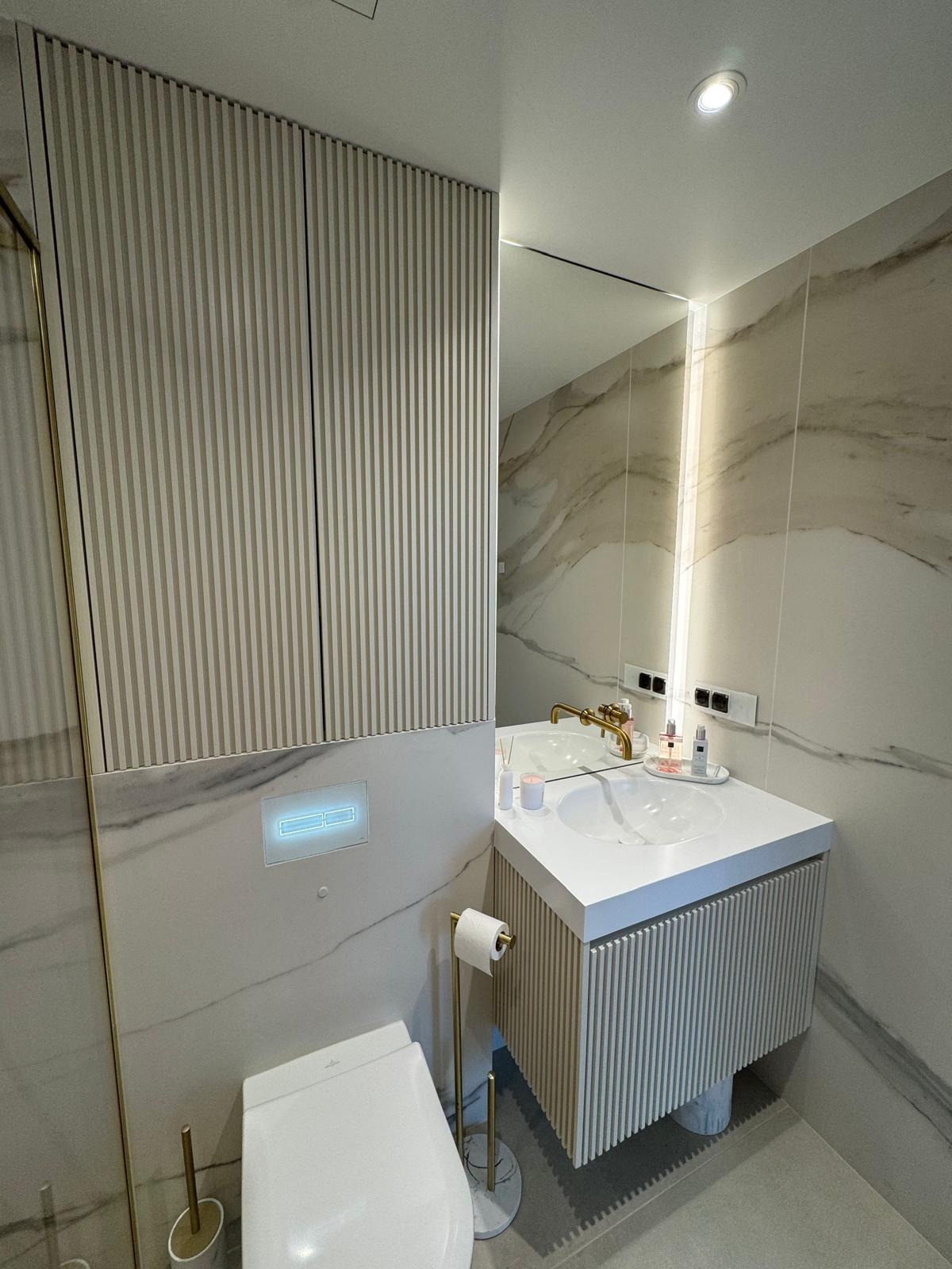 Nowoczesna łazienka z szafką pod umywalką i szafą wnękową z lamelami w odcieniach beżu i bieli, marmurowe płytki, złota armatura i akcesoria.