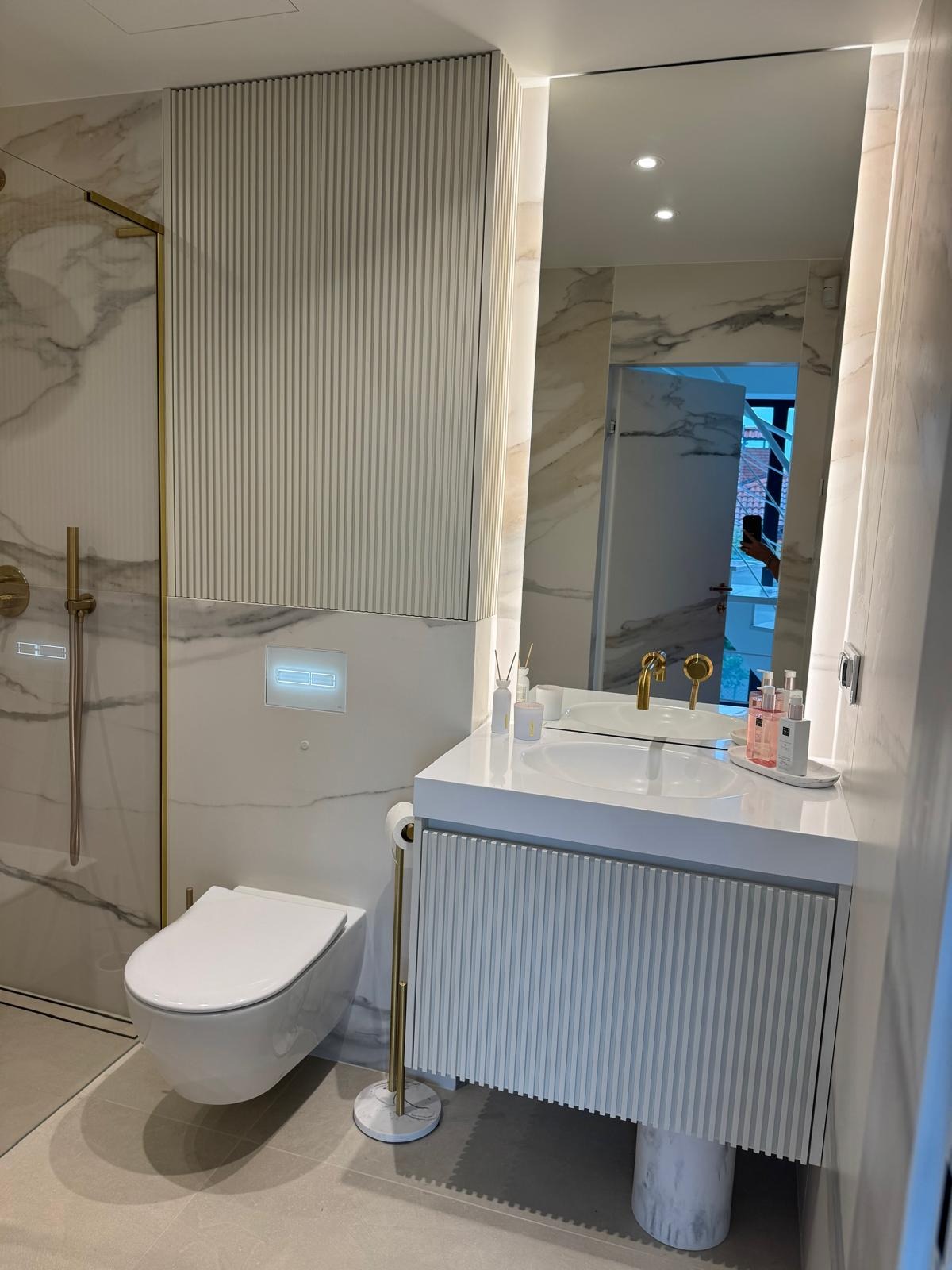 Nowoczesna łazienka z marmurowymi płytkami, białym blatem umywalki z ryflowanym frontem i podświetlanym lustrem. Złote akcenty dodają elegancji.