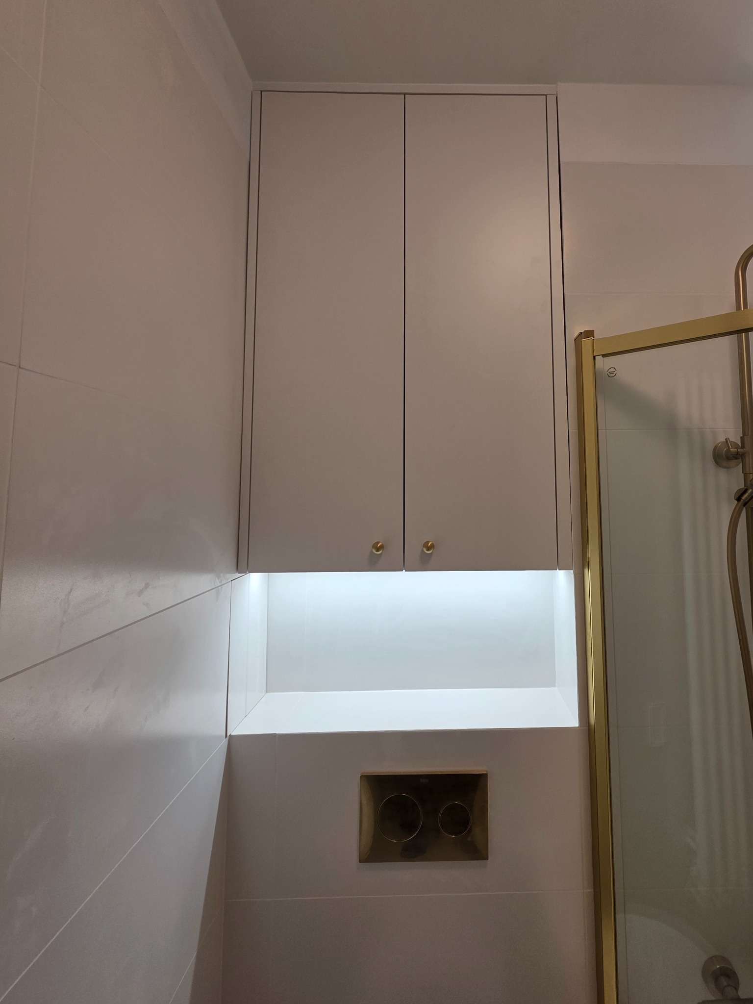 Biała szafka łazienkowa pod sufitem z oświetleniem LED nad złotym przyciskiem spłuczki, obok kabiny prysznicowej ze złotymi ramami.