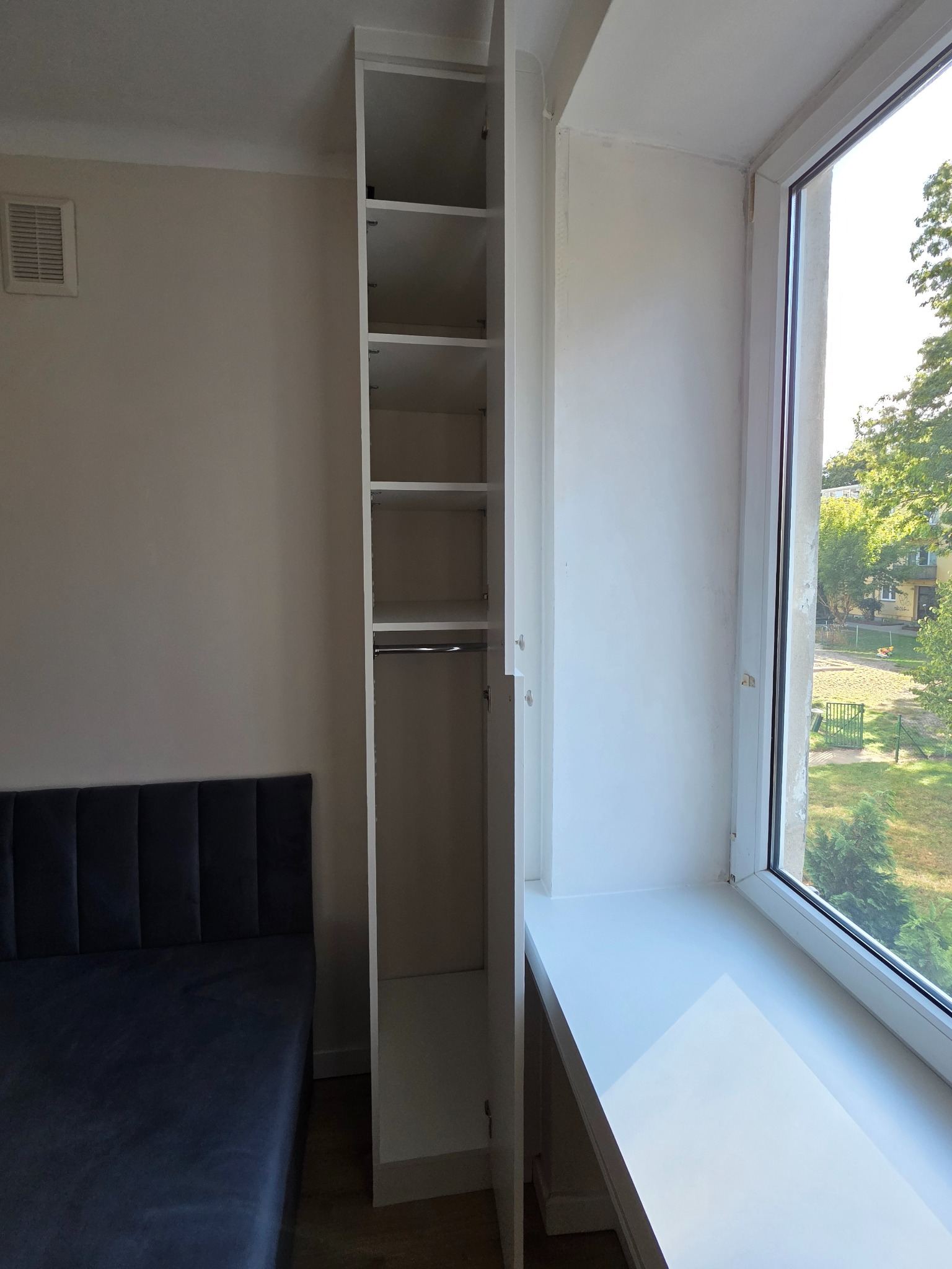 Wysoka, wąska, biała szafa wnękowa z otwartymi drzwiami, z półkami i drążkiem na ubrania, obok okna z szerokim parapetem. Widok na zieleń za oknem.