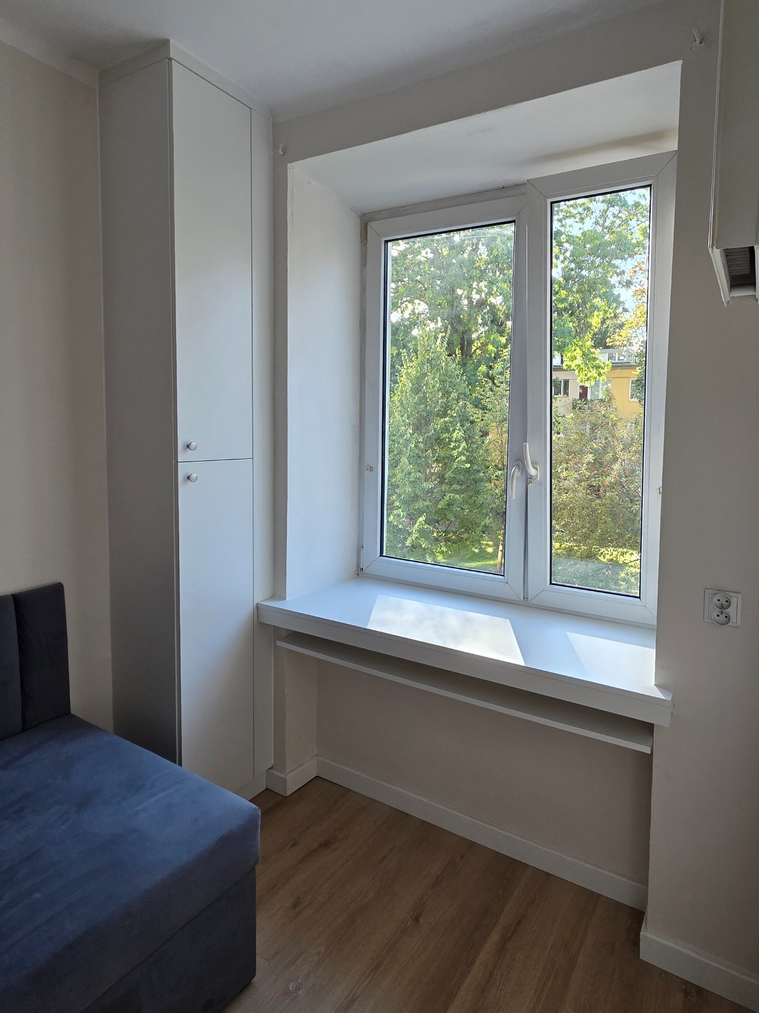 Biała szafa wnękowa z dwoma segmentami, dopasowana do okna z szerokim parapetem, drewniana podłoga. Widok na zieleń za oknem.