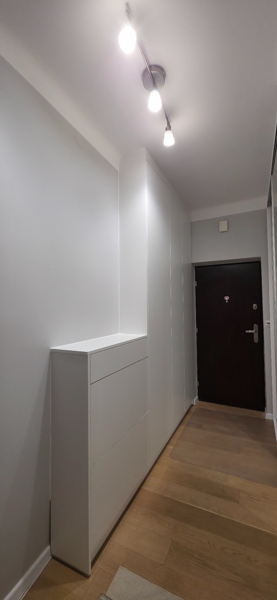 Biała szafa wnękowa z szufladami, dopasowana do przedpokoju. Minimalistyczny design, drewniana podłoga, oświetlenie sufitowe.