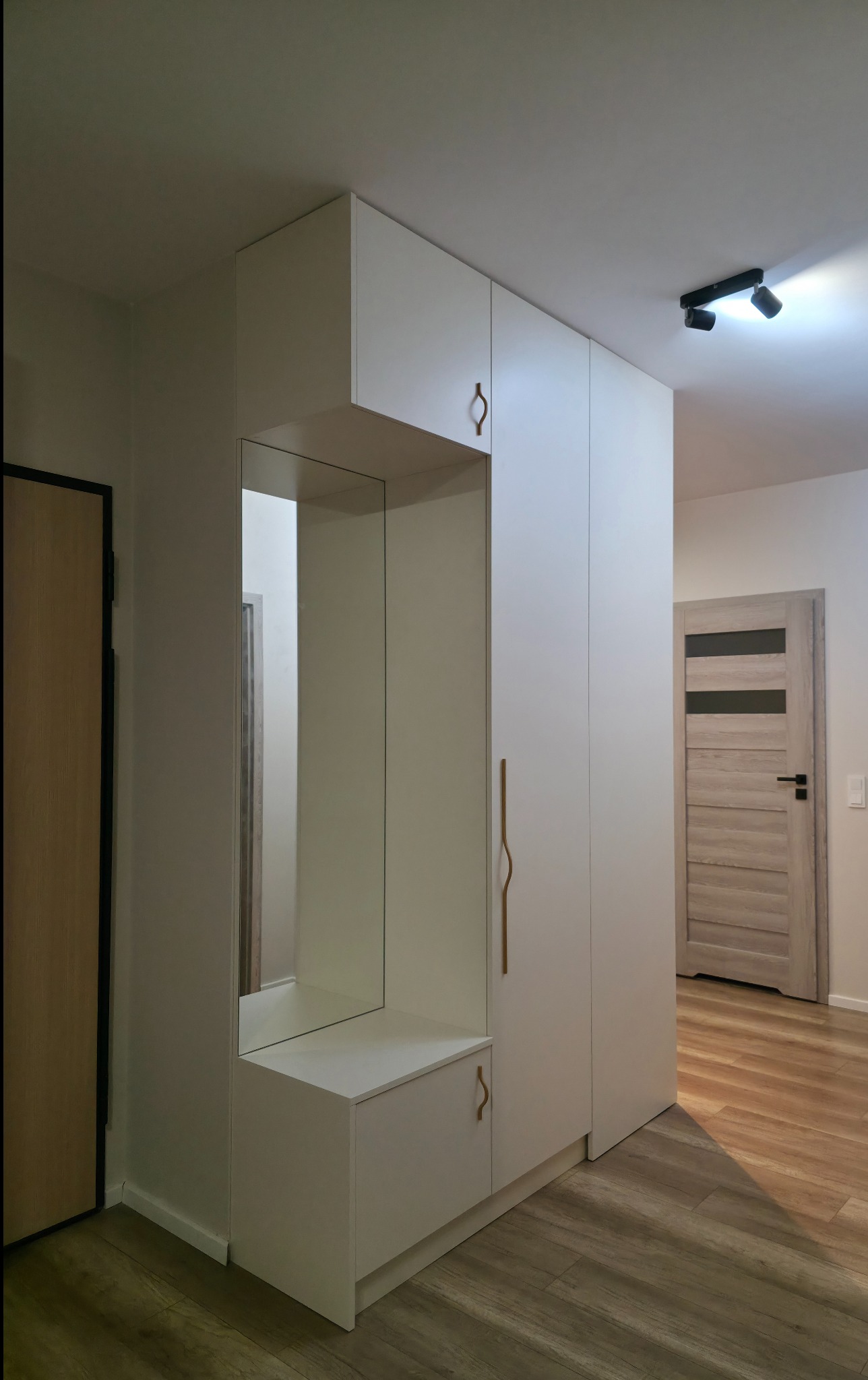 Biała szafa wnękowa z lustrem i siedziskiem w przedpokoju. Minimalistyczny design, drewniana podłoga, nowoczesne oświetlenie sufitowe.