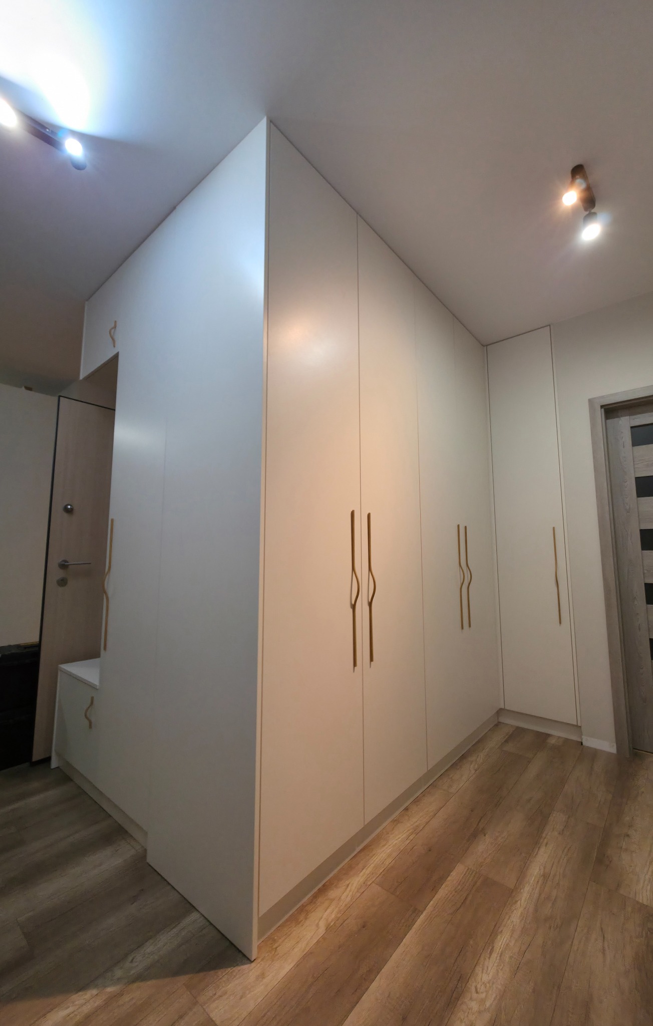 Biała szafa wnękowa z drewnianymi uchwytami w przedpokoju, widok z dołu, oświetlenie punktowe, panele podłogowe imitujące drewno. Estetyczne wykończenie.