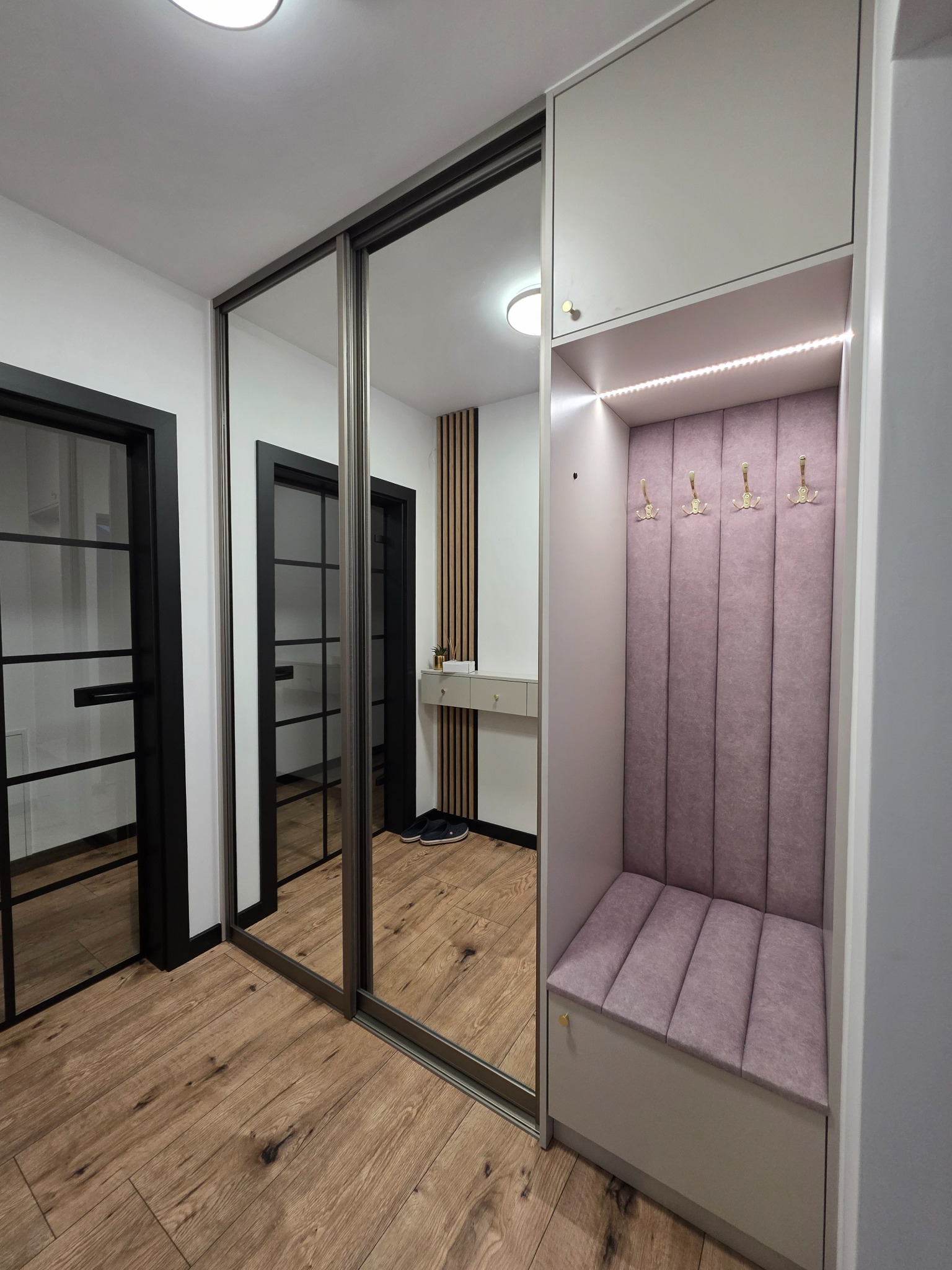 Nowoczesny przedpokój z szafą wnękową z lustrzanymi drzwiami przesuwnymi i siedziskiem z wieszakami, oświetlony ledami. Widoczna podłoga z paneli i drzwi.