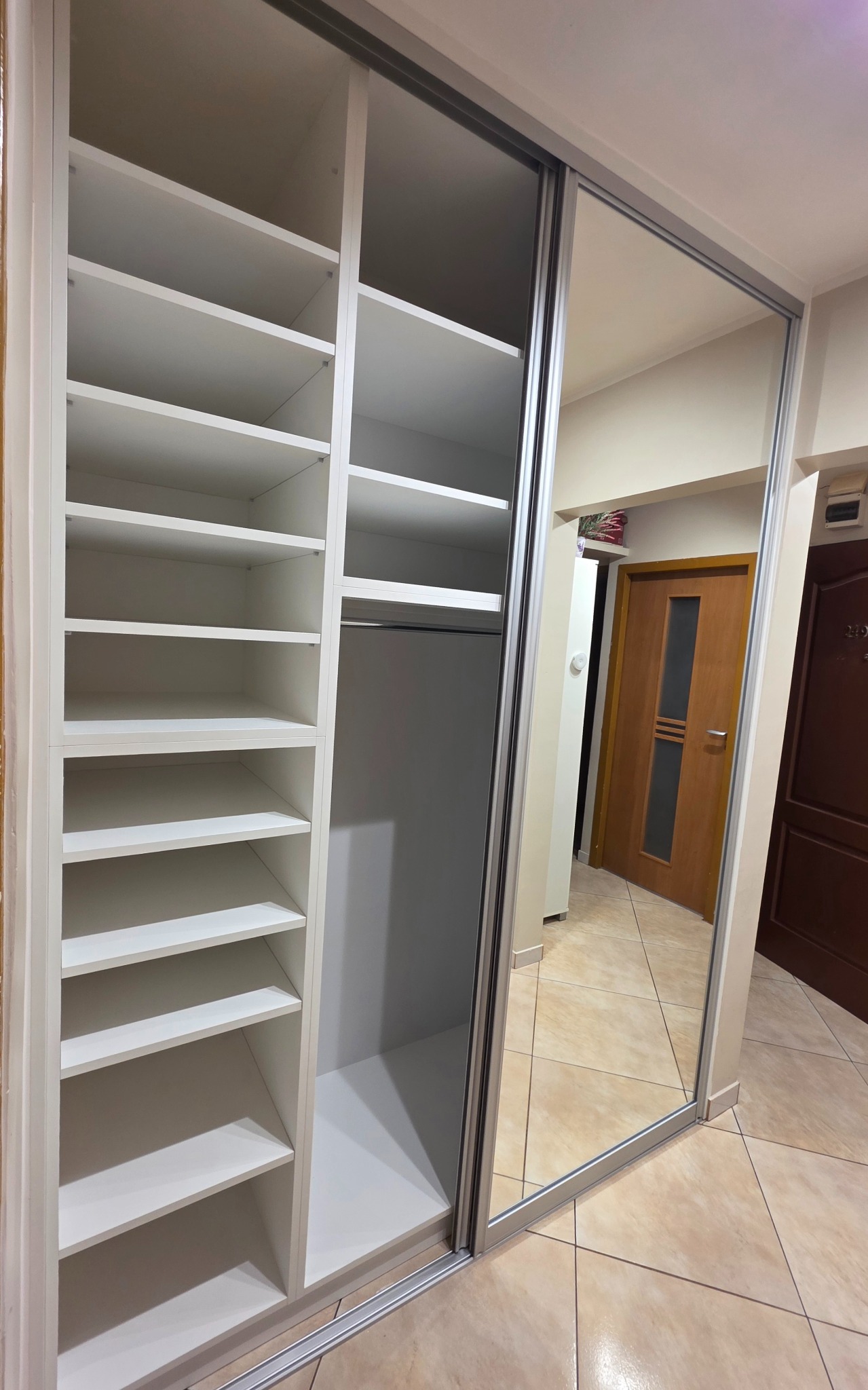 Biała szafa wnękowa z przesuwnymi drzwiami lustrzanymi, widoczne półki i drążek na ubrania. W lustrze odbija się fragment korytarza z drzwiami.