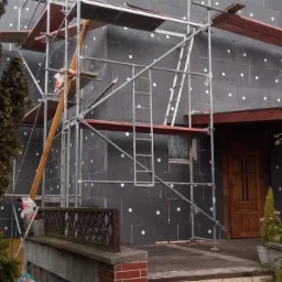 Dom w trakcie prac: szara elewacja z widocznym ociepleniem, białe kołki, rusztowanie metalowe, drewniane deski na rusztowaniu, wejście do domu z ceglanym wykończeniem, worki na śmieci i niebieskie...