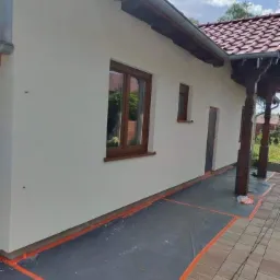 Świeżo pomalowana, jasna elewacja domu jednorodzinnego z brązowymi oknami, zabezpieczona folią malarską na kostce brukowej. Widoczne ślady farby na folii.