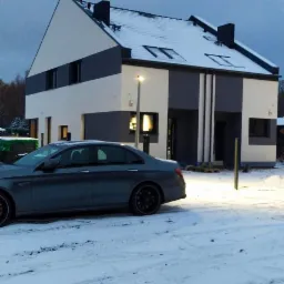 Nowoczesny dom jednorodzinny z biało-szarą elewacją, pokryty śniegiem, z szarym samochodem zaparkowanym na ośnieżonym podjeździe, oświetlony przez latarnię uliczną.