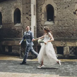Uśmiechnięta para młoda biegnie trzymając się za ręce na tle ceglanej ściany z oknami w kształcie łuków; panna młoda w koronie i z bukietem w ręku.