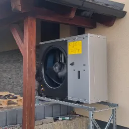 Szara pompa ciepła zamontowana na metalowym stelażu przy ścianie budynku, widoczne narzędzia i paleta w tle.