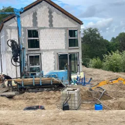 Prace ziemne przy budowie domu: niebieska wiertnica na gąsienicach, żółta mini koparka JCB i pracownik w wykopie, widoczny budynek w stanie surowym otwartym z wstawionymi oknami.