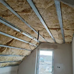 Izolacja poddasza wełną mineralną pomiędzy metalowymi profilami, widoczne okno z widokiem na zewnątrz.