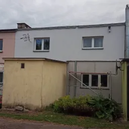 Dwukondygnacyjny budynek z szarą elewacją, anteną satelitarną i rurą spustową, widoczny z zewnątrz, z ż&oacute;łtą przybud&oacute;wką i roślinnością u podstawy.