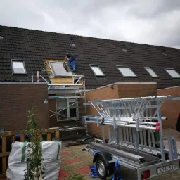 Montaż okna dachowego na tle pochmurnego nieba, widoczne rusztowanie, przyczepka z drabinami oraz narzędzia na dachu.