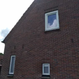 Czerwona ceglana ściana domu z trzema oknami PCV o różnych rozmiarach i kształtach, z odbiciem chmur na górnym oknie.