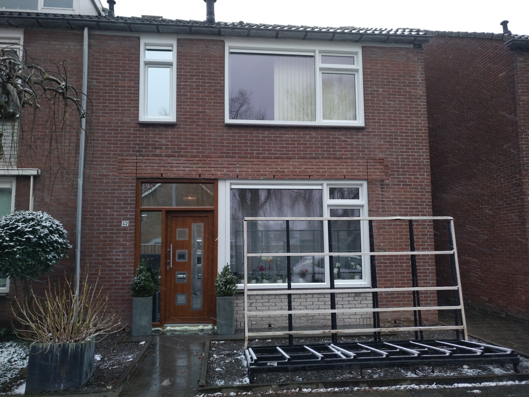Brązowy, dwukondygnacyjny dom z cegły z białymi ramami okiennymi i drewnianymi drzwiami wejściowymi. Na pierwszym planie widoczny stojak na okna/szyby pokryty śniegiem i donice z roślinami po obu...