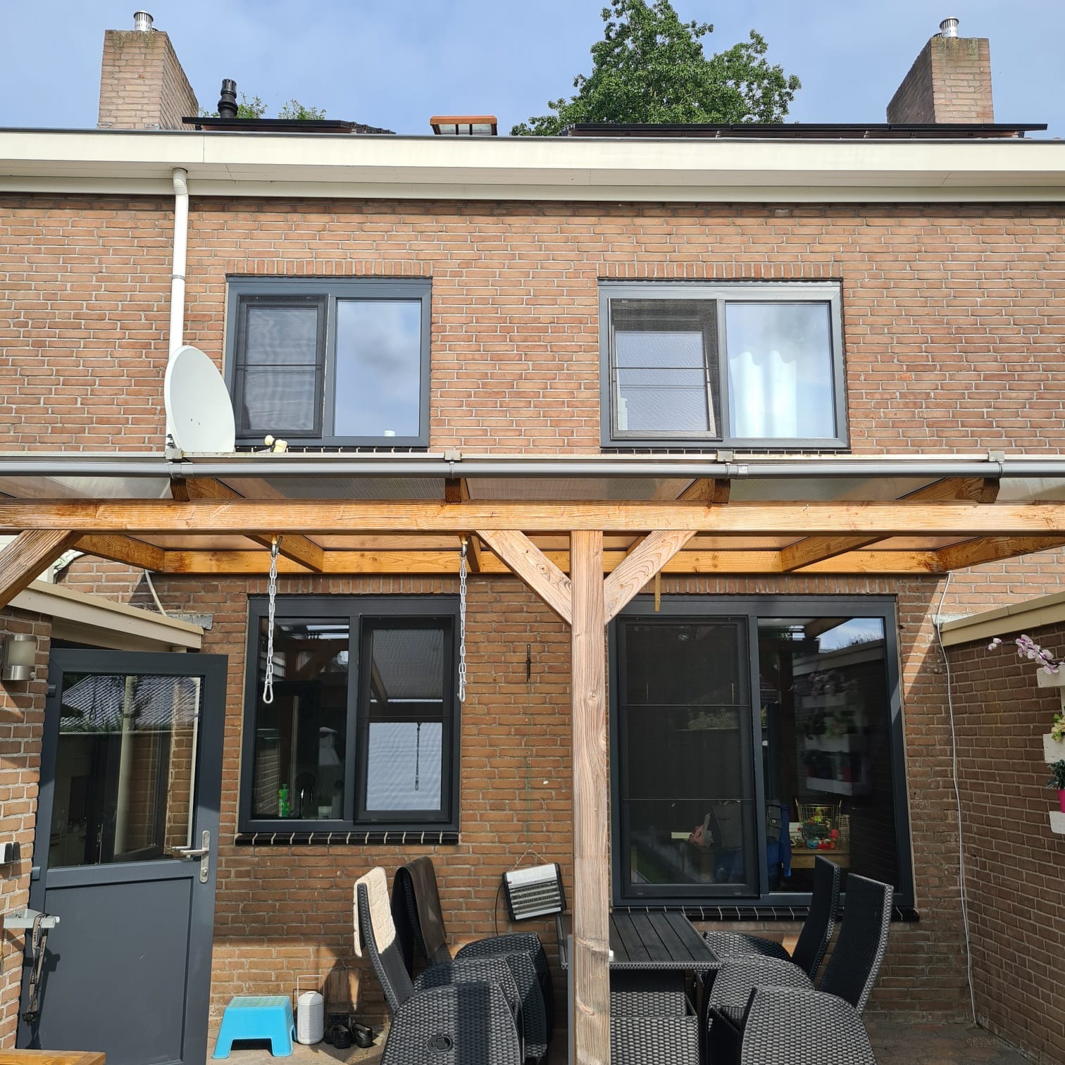 Widok na elewację domu z cegły z zewnątrz, z zamontowanymi oknami z czarnymi ramami i drzwiami tarasowymi, widoczny drewniany zadaszony taras z meblami ogrodowymi.