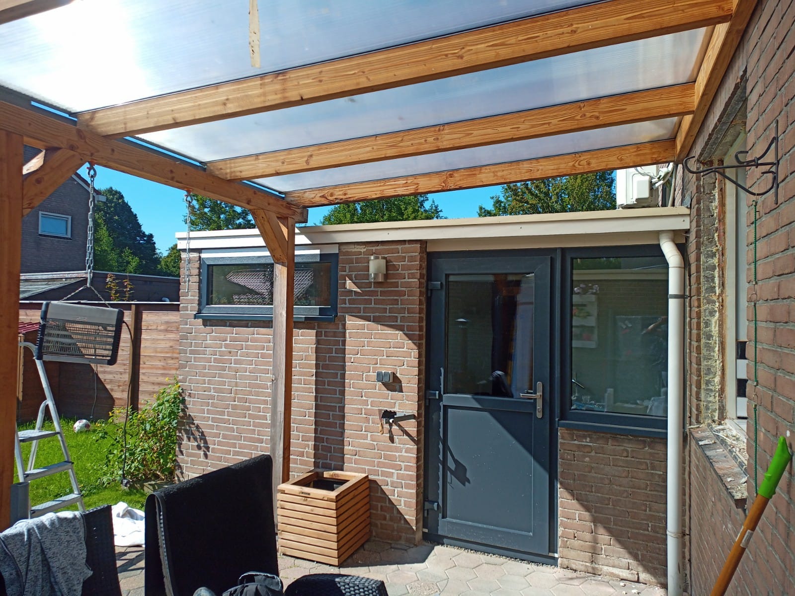 Zadaszony taras z drewnianą konstrukcją i przezroczystym poliwęglanowym dachem, widoczna szara furtka z oknem, ceglana ściana budynku oraz drabina oparta o drewniany słup.