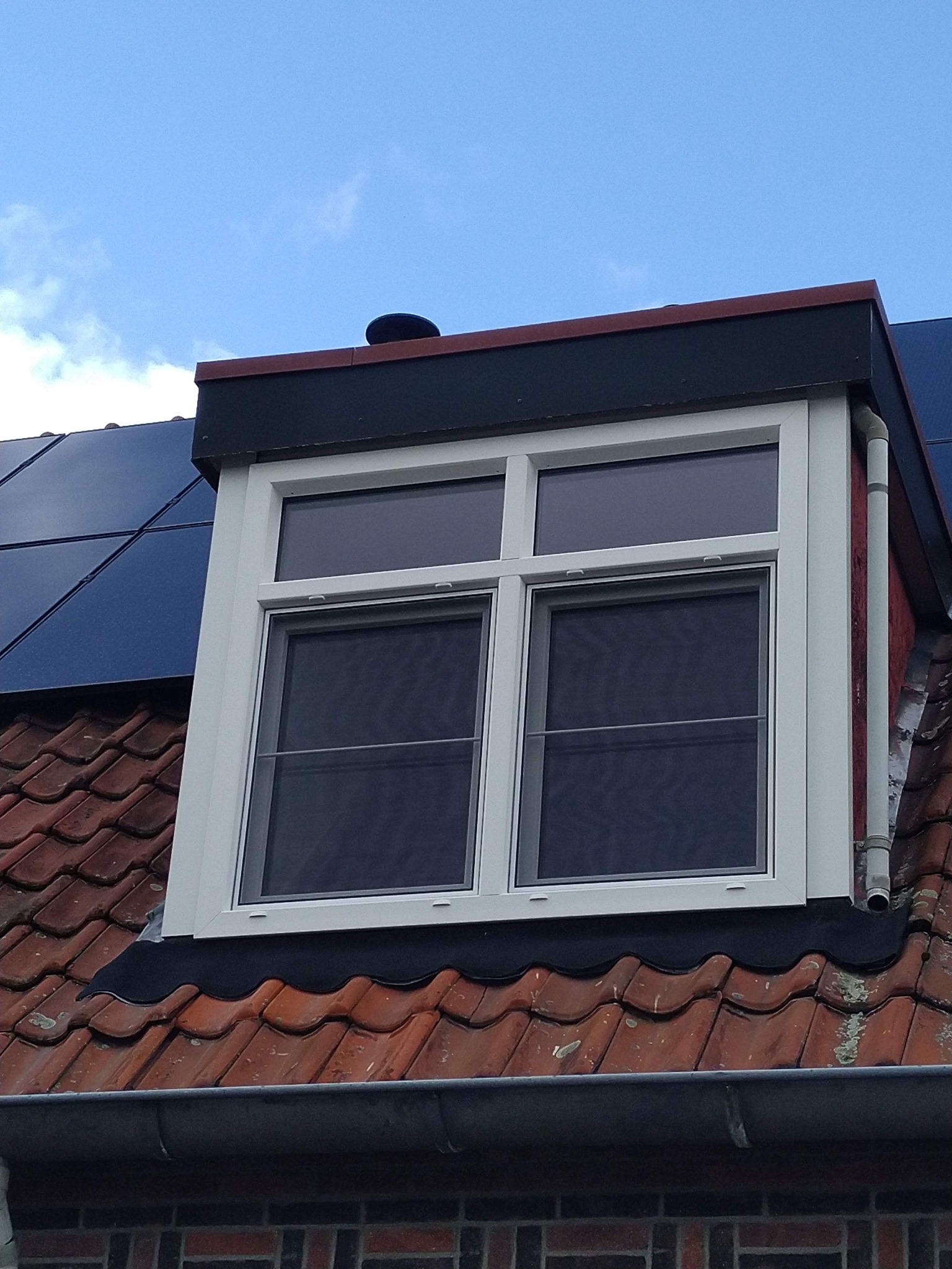 Białe okno dachowe z moskitierą, zamontowane na czerwonej dachówce, widoczne panele słoneczne i rynna.