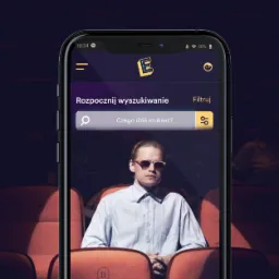 Ekran smartfona prezentujący interfejs aplikacji E-kino z motywem kinowym, gdzie w tle widoczny jest mężczyzna w okularach siedzący na fotelu kinowym. Aplikacja umożliwia wyszukiwanie i kupowanie...