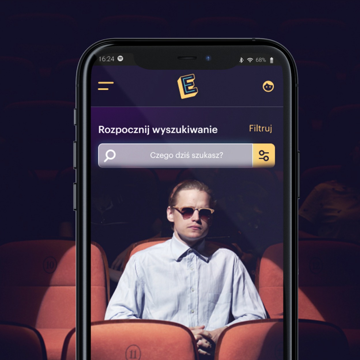 Ekran smartfona prezentujący interfejs aplikacji E-kino z motywem kinowym, gdzie w tle widoczny jest mężczyzna w okularach siedzący na fotelu kinowym. Aplikacja umożliwia wyszukiwanie i kupowanie...
