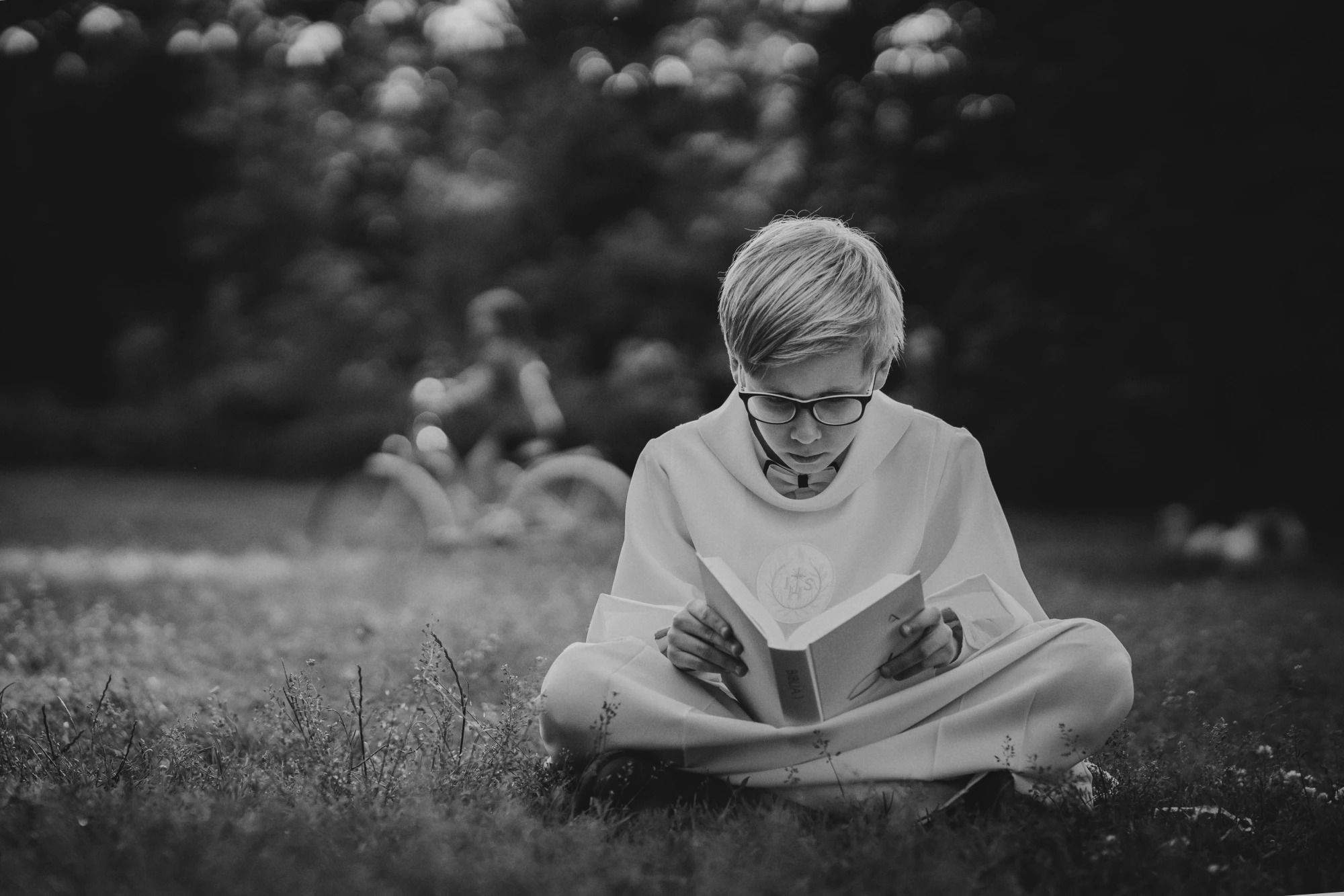 Chłopiec w stroju komunijnym, siedzący na trawie i czytający książkę, w tle rozmyty rower