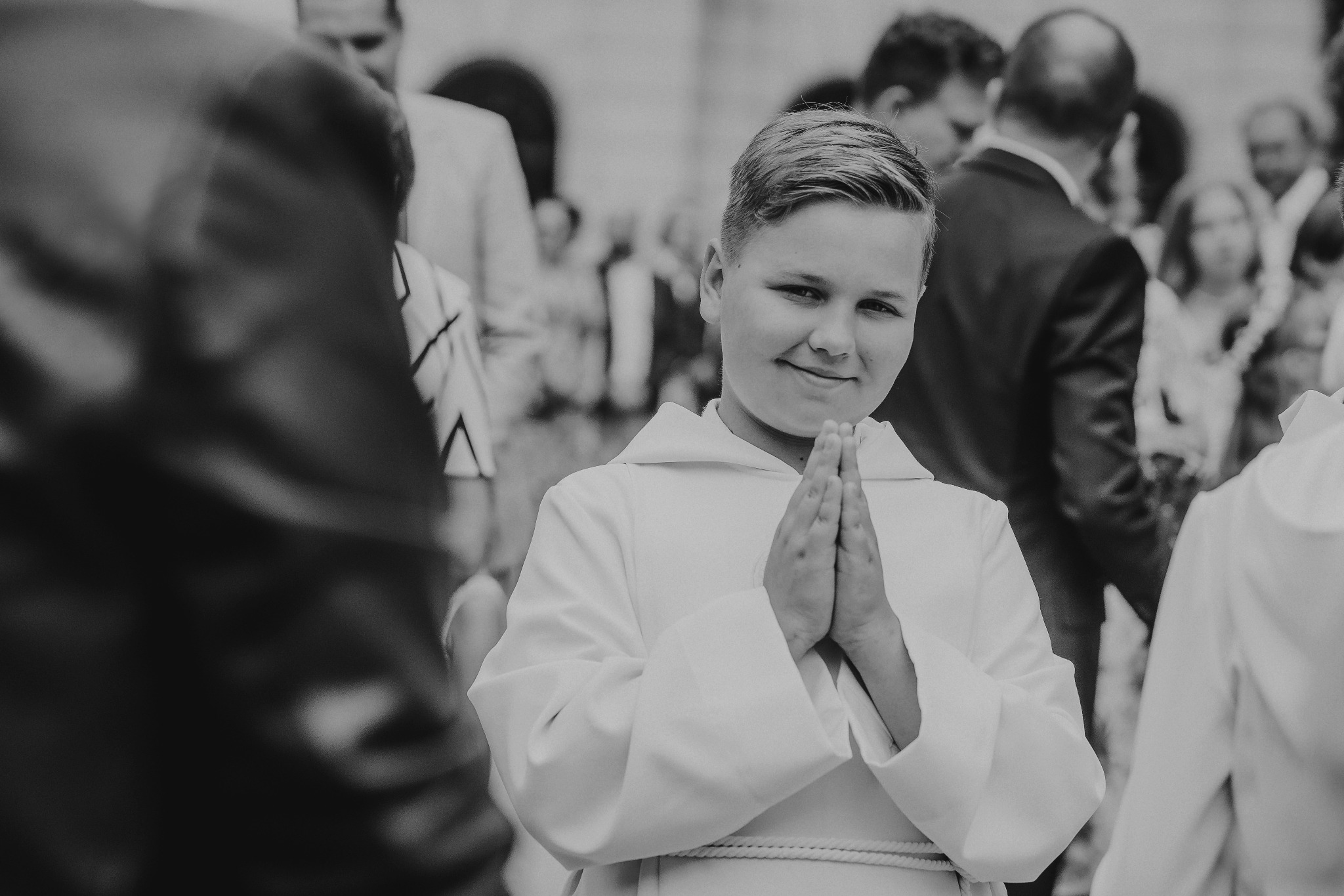 Chłopiec w białej albie, ze złożonymi dłońmi, uśmiecha się na tle rozmytych postaci podczas uroczystości.