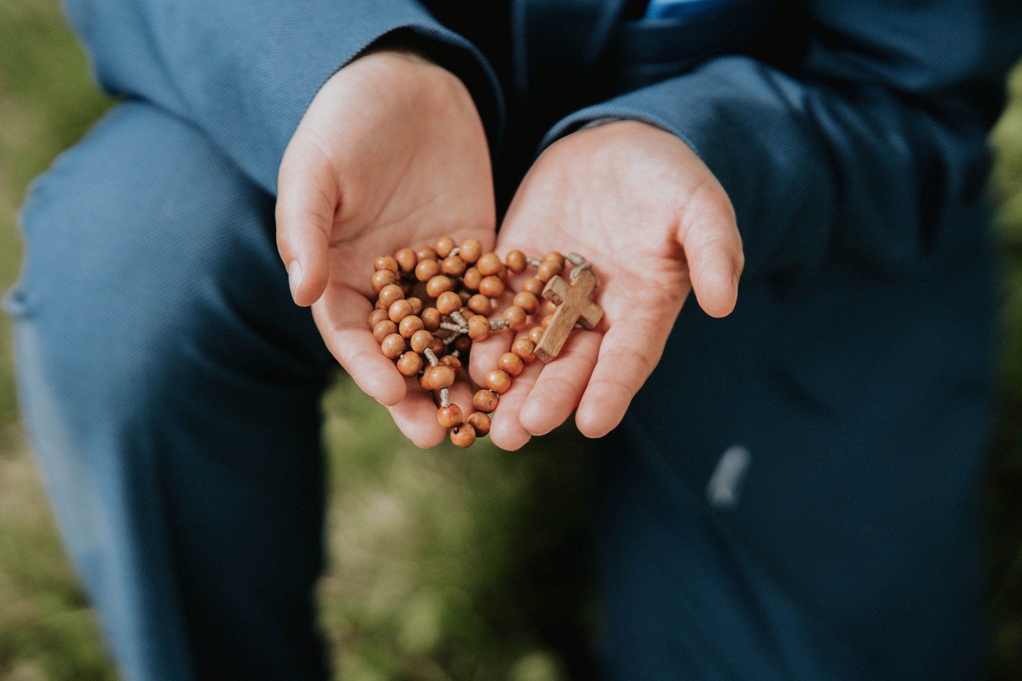 Dłonie w niebieskim garniturze trzymają drewniany różaniec z krzyżykiem na zewnątrz.
