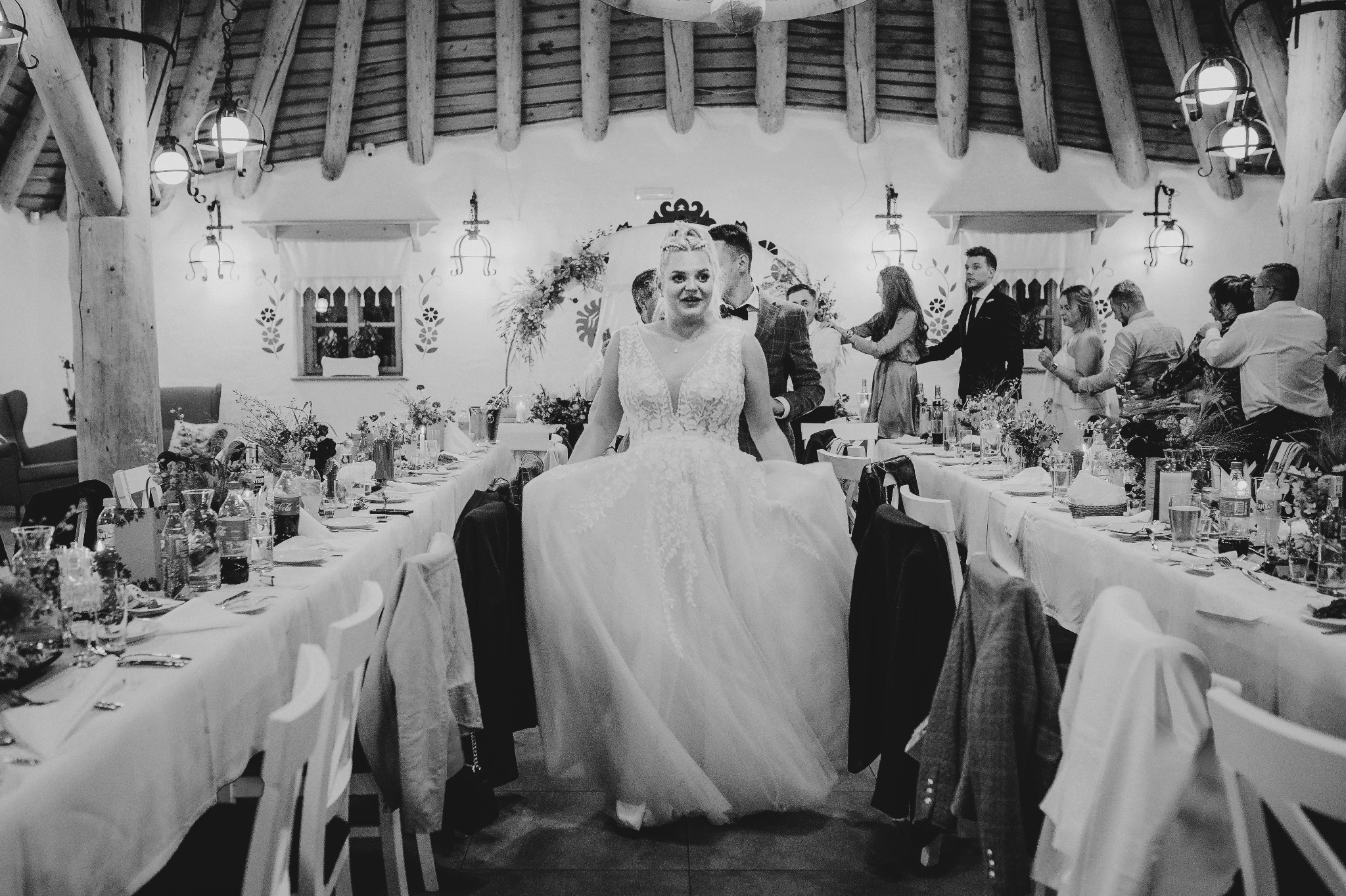 Panna młoda w koronkowej sukni spaceruje między stołami weselnymi w rustykalnej sali, goście w tle, czarno-biała fotografia.