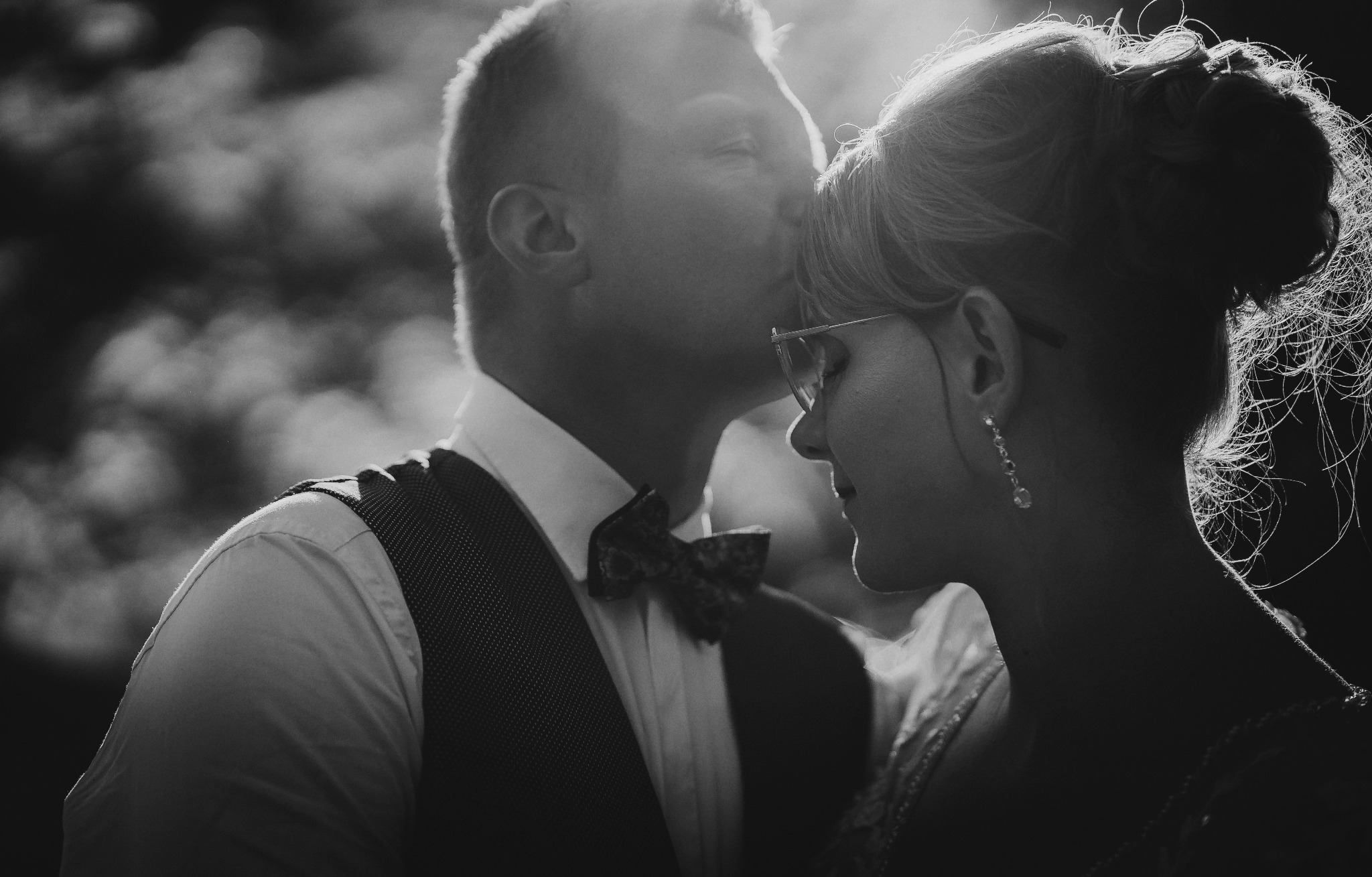 Czarno-białe zdjęcie pary młodej: pan młody w muszce i kamizelce całuje pannę młodą w czoło, panna młoda w okularach i z upiętymi włosami.