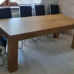 Prostokątny stół z drewna w jasnym odcieniu, otoczony krzesłami z czarnymi siedziskami i metalowymi nogami w nowoczesnym wnętrzu z jasną podłogą i dekoracyjną ścianą z kamienia.