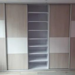 Szafa przesuwna z jasnego drewna z białymi wstawkami, z otwartą częścią środkową z półkami, na tle różowej ściany.