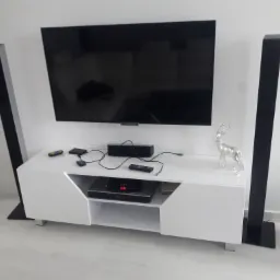 Biała szafka RTV z czarnymi głośnikami kolumnowymi po bokach, telewizorem na ścianie i srebrną figurką jelenia jako dekoracją.