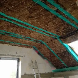 Poddasze w trakcie izolacji termicznej: wełna mineralna zabezpieczona sznurkiem i zieloną folią, widoczne krokwie i fragment ściany z oknem.