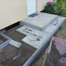 Montaż kostki brukowej na schodach wejściowych do domu, widoczne poziomice, młotek, kielnia i taczka z piaskiem.