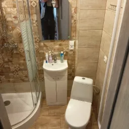 Ukończona łazienka z kabiną prysznicową, umywalką z szafką, toaletą i lustrem, wykończona płytkami w odcieniach beżu i brązu; w lustrze odbija się osoba w kombinezonie roboczym.