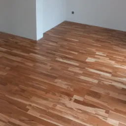 Nowo ułożona, parkietowa podłoga z jasnego drewna w pustym, pomalowanym na biało pokoju, widoczny fragment gniazdka elektrycznego przy podłodze.