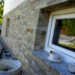 Fragment elewacji budynku wykończonej dekoracyjnym kamieniem w odcieniach beżu i szarości, z widocznym oknem i parapetem z granitu oraz wiadrem z zaprawą murarską sugerującym trwające prace...