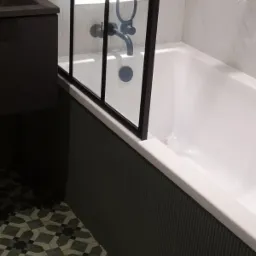 Nowoczesna łazienka z białą wanną, czarną armaturą i geometryczną podłogą; szklany parawan nawannowy z czarnymi szprosami.