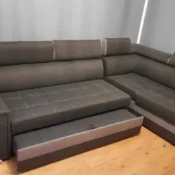 Rozłożona narożna kanapa w odcieniach szarości z wysuwanym pojemnikiem na pościel, widoczne drewniane panele podłogowe oraz dekoracja ścienna z imieniem Igor.