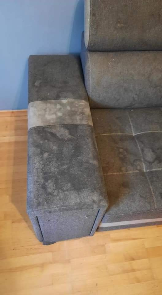 Fragment brudnej, szarej kanapy z plamami na tapicerce, widoczny bok i część siedziska na drewnianej podłodze.