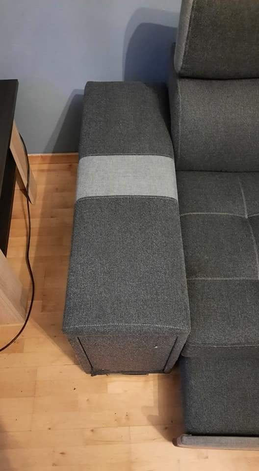 Fragment szarej sofy z podłokietnikiem z dwukolorowej tkaniny, widoczny fragment drewnianej podłogi i stolika.