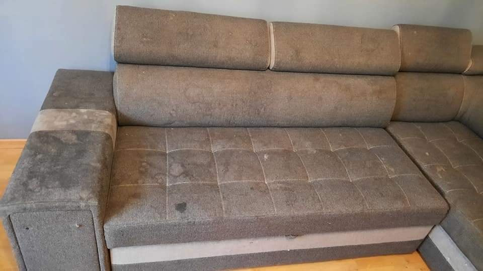 Brudna, narożna kanapa z tapicerką w odcieniach szarości, widoczne plamy i zabrudzenia na siedzisku, oparciu i podłokietnikach.