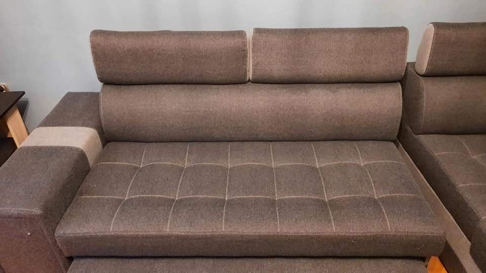 Szary narożnik z podnoszonymi zagłówkami i pikowanym siedziskiem, widoczny fragment stolika z jasnego drewna w tle.